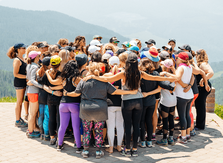 A large group of women standing in a circle outdoors, arms around each other.