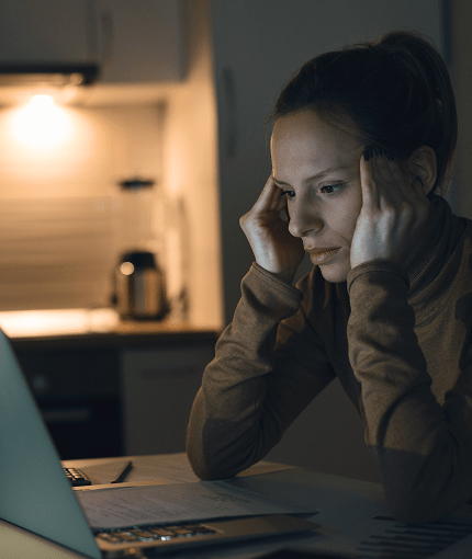 A woman sitting alone at her kitchen table late at night, head in her hands over an open laptop.